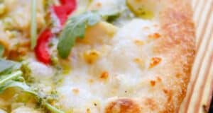 Close-up of a pizza in one of the ten dining recommendations for your Disney vacay, featuring a golden-brown crust, melted cheese, sliced red peppers, vibrant green arugula leaves, and a drizzle of green pesto sauce on top.
