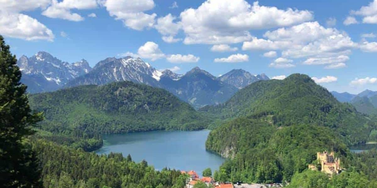 Scenic view of a lush green valley with a lake surrounded by forested hills and snow-capped mountains under a partly cloudy blue sky. A small village with red-roofed buildings, reminiscent of one you might find if you plan a Disney-inspired trip to Germany, is visible near the lake.