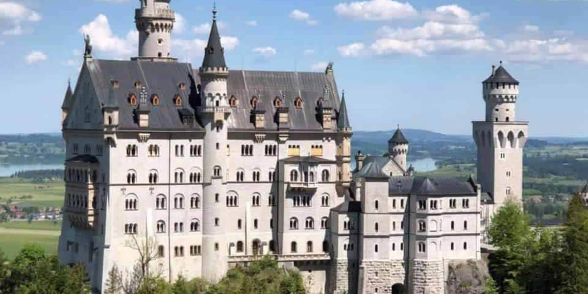 A large, ornate castle with numerous towers and spires stands atop a hill, reminiscent of a Disney fairy tale. The lush landscape of trees and a distant body of water creates an enchanting backdrop. The sky is clear with a few scattered clouds—perfect for planning your Disney-inspired trip to Germany.