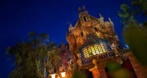 A gothic-style mansion with ornate architecture and glowing windows is illuminated against a dark night sky. The building, which holds Disney's Haunted Mansion ride, is surrounded by trees, adding to the eerie atmosphere.