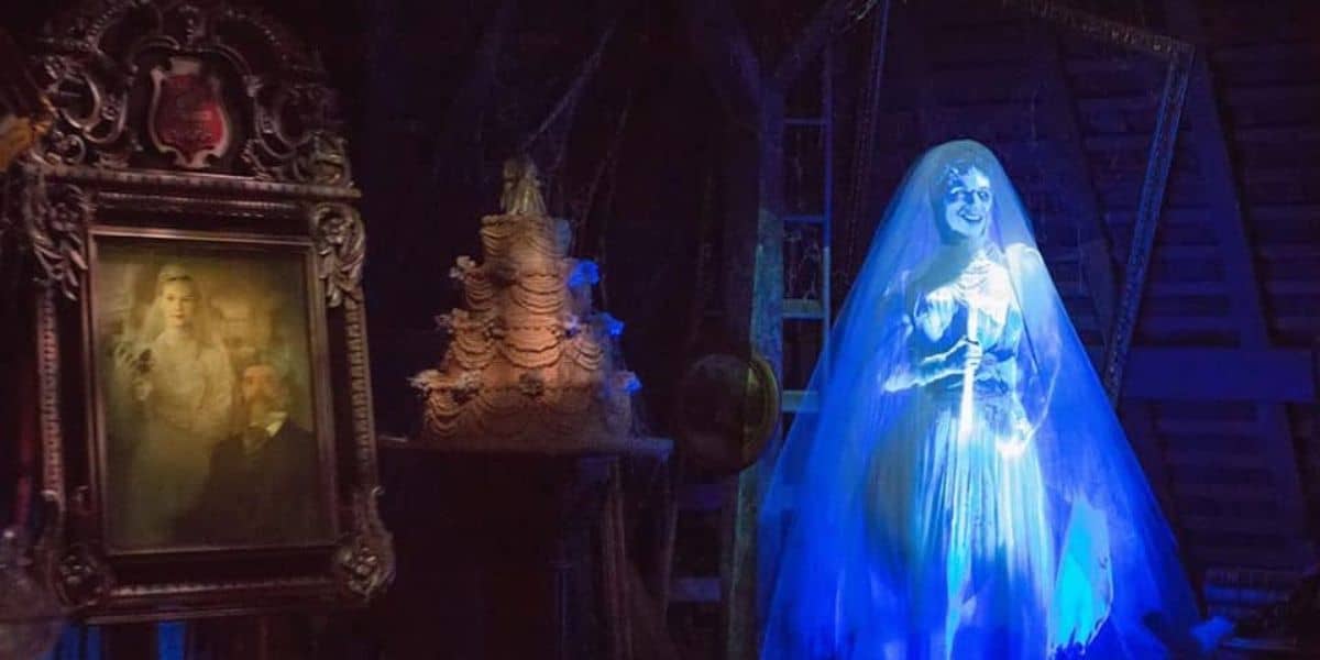 A ghostly figure of a bride in an illuminated white gown and veil stands beside a multi-tiered wedding cake in a dark, eerie room reminiscent of Disney's Haunted Mansion ride. A framed, aged portrait of a couple hangs on the wall to the left. The room's atmosphere is otherworldly and haunted.