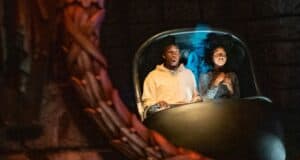 A man and woman ride in a dark amusement park attraction at Disney's Haunted Mansion, sitting in a two-seater pod. The man looks surprised with his mouth open, while the woman appears focused on the ride. The scene is dimly lit, creating a suspenseful atmosphere.