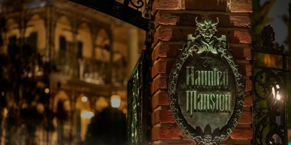 A nighttime image of a brick pillar with an ornate metal sign reading "Haunted Mansion," featuring a demonic face at the top. The background shows a dimly lit, gothic-style building with intricate ironwork, adding to the eerie atmosphere reminiscent of Disney's Haunted Mansion ride.