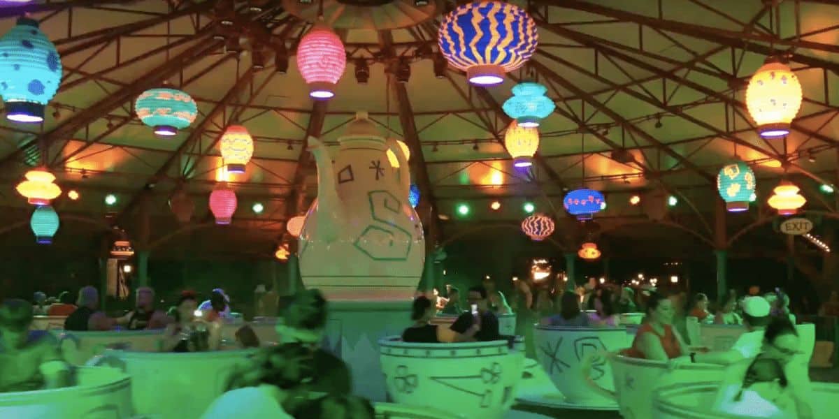 People are enjoying a tea cup ride inside a brightly-lit, colorful indoor amusement park attraction. The ride is decorated with vibrant lantern-shaped lights hanging from the ceiling and a large teapot in the center. Despite the lively and fun atmosphere, this is one of the rides to skip at Disney World.