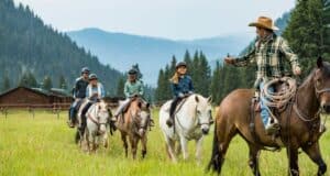 A group of people enjoy a thrilling horseback ride through a lush green field with mountains in the backdrop, guided by an expert. They're equipped with helmets and casual clothing, just like an adventure from New Vacations by Disney. In the distance, a charming wooden cabin can be seen.