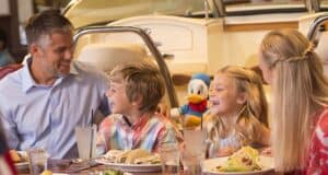 A family of four, including two children, enjoys a meal at a restaurant. They are smiling and sitting at a table with food and drinks. A plush toy resembling a cartoon duck is visible in the background, hinting at the delightful dining options at Disney World.