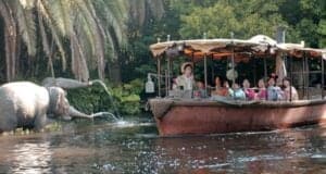 A group of people enjoy a jungle-themed boat ride, reminiscent of a classic Disney ride, passing a lifelike elephant statue spraying water near lush greenery. The boat is covered and steered by a guide, creating a playful safari atmosphere that embodies an exciting change from the ordinary.