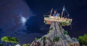 A dramatic night sky with a partially cloud-covered moon illuminates a ship structure perched atop a rocky formation, reminiscent of Disney Typhoon Lagoon. Palm trees and scattered wooden crates decorate the scene below, evoking the adventure of a volcanic island.