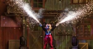 Mickey Mouse, dressed as a magician, stands with arms raised, shooting sparkling fireworks in front of a rustic wooden building. The scene captures the energetic and magical atmosphere reminiscent of a classic Disney Nighttime Show.
