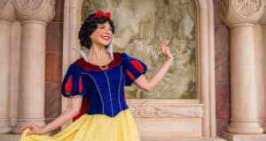 A person dressed as a fairytale character stands in front of a decorative backdrop featuring a castle at Disney World Princess Week. They wear a red bow, blue and red top, and yellow skirt, posing with one hand raised. The setting is ornate with stonework.