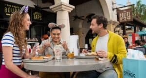 Three people sit at an outdoor restaurant table, smiling and enjoying a meal in a festive atmosphere. One wears mouse-ear headbands, hinting at Disney fun. In the background, a sign for "The Polite Pig" adds to the lively setting, making it an ideal spot for any Disney guest to unwind.