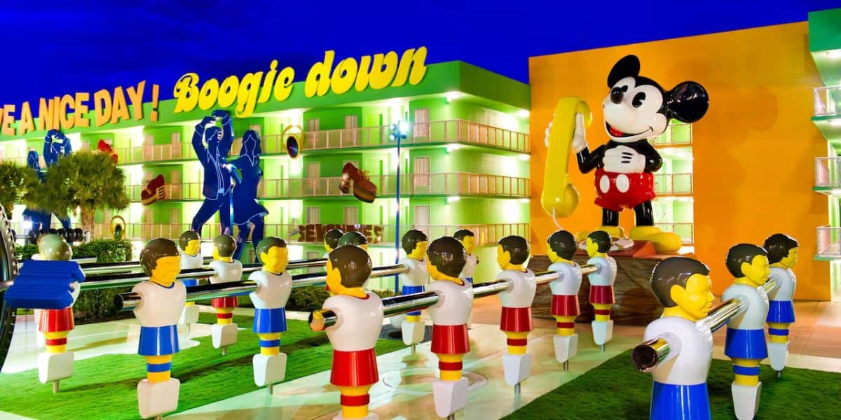 The vibrant exterior of the Disney Resort features large statues of a smiling mouse in a yellow outfit holding a phone, alongside a giant foosball table. Bright signs proudly display "Dance Da..." and "Boogie Down!" against the backdrop of a vivid blue sky.