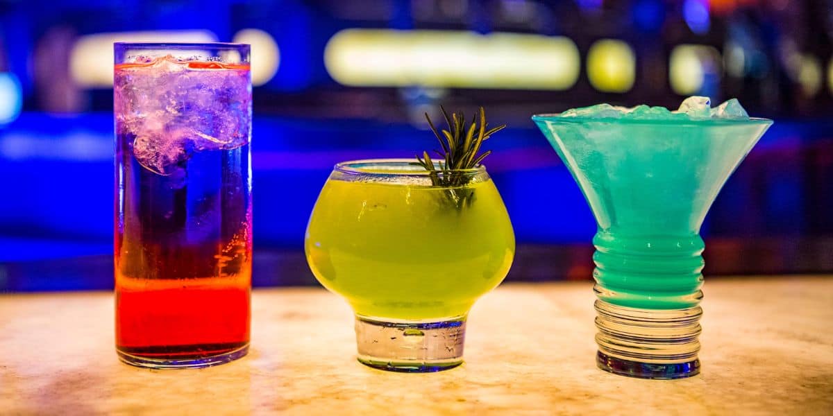 Three colorful cocktails on a bar counter: a tall glass with a red drink and ice, a round glass with a green drink garnished with rosemary, and Disney World's frosty blue signature cocktail in a wide-mouthed glass. A dim, blue-lit background enhances the scene.