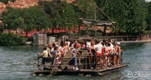 A group of people stand closely together on a wooden raft, floating on a calm body of water. Trees and a small structure reminiscent of the Magic Kingdom are visible in the background. The scene appears lively under the clear weather, with a whimsical air that hints at an adventure worthy of any bear's tale.