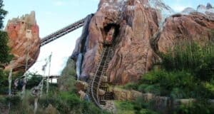 A roller coaster track emerges from a large mountain, designed to resemble Expedition Everest with a rugged peak and cave-like opening. The track, filled with passengers, descends steeply, surrounded by lush greenery and a blue sky above. Various structures decorate the mountainside.