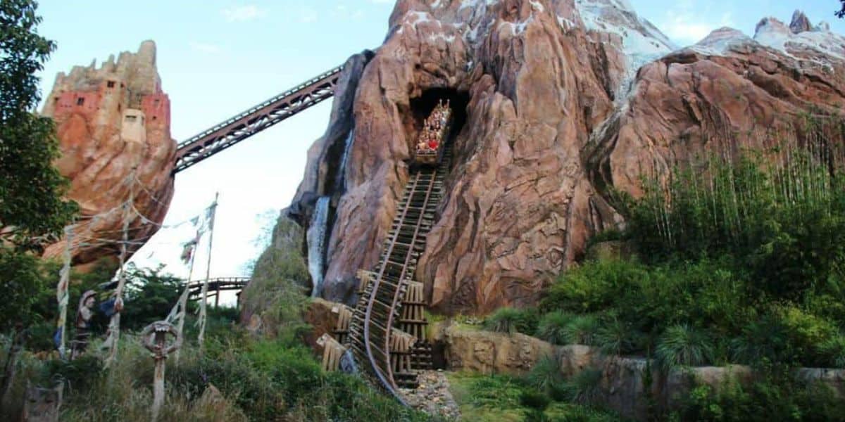 A roller coaster track emerges from a large mountain, designed to resemble Expedition Everest with a rugged peak and cave-like opening. The track, filled with passengers, descends steeply, surrounded by lush greenery and a blue sky above. Various structures decorate the mountainside.
