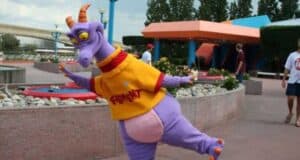 A purple dragon character, reminiscent of Disney changes, wears an orange and yellow shirt and stands playfully on one leg in an outdoor area. With orange horns and a cheerful expression, the lively scene includes people casually walking by planters and colorful structures.