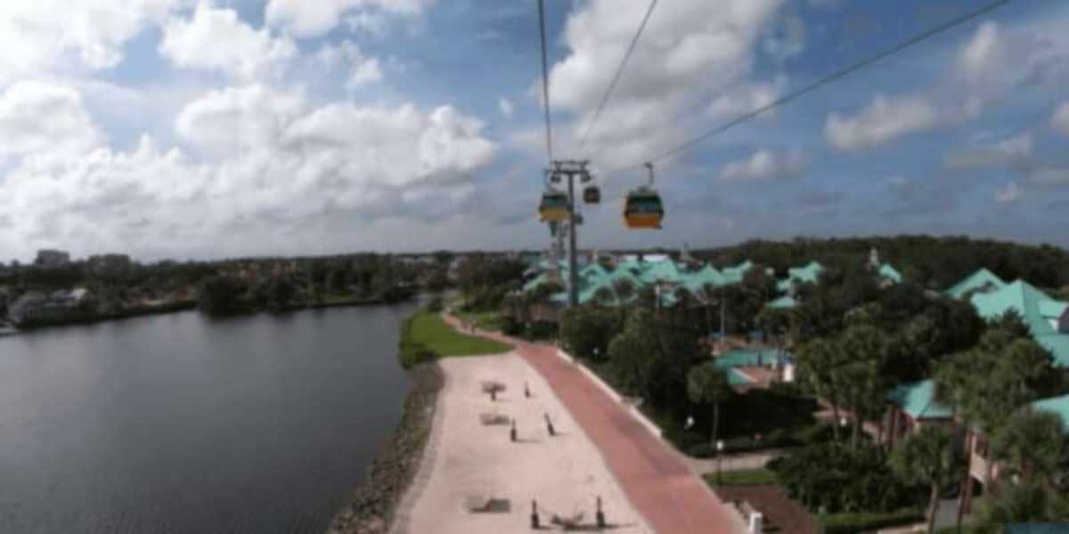 Experience a Disney World vacation with a scenic view of a cable car system gliding over a tree-lined resort area, featuring turquoise-roofed buildings. Marvel at the tranquil river on the left and enjoy the charming pathway below under a partly cloudy sky.