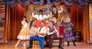 A group of six performers in colorful, western-style costumes pose on stage at Disney World. They hold props and smile, with a backdrop of festive decorations and the words "Hoop-Dee-Doo" visible. The lively, theatrical atmosphere hints at the fun waiting just beyond the stage, perhaps with a splash of booze for the adults.