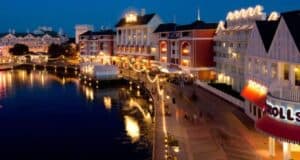 Nighttime scene of a lively waterfront boardwalk with colorful buildings, bright lights reflecting on the water, and people strolling along the path. The area has a festive atmosphere, featuring various shops and restaurants.