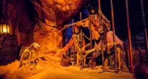 Skeletons dressed as pirates behind jail bars extend a bone to a dog with keys in its mouth. The dimly lit scene, reminiscent of a Disney adventure, unfolds in a cave-like setting with an air that echoes the whimsical charm of Country Bears.