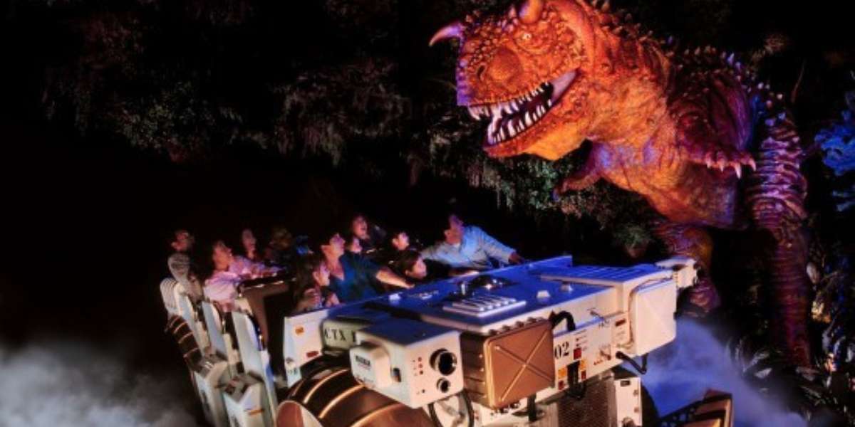 A group of people in a ride vehicle react excitedly as they encounter a large, animatronic dinosaur with sharp teeth in a dark, themed attraction setting. The atmosphere is enhanced with smoke effects and dramatic lighting.