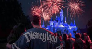 A couple stands in front of a crowd watching a vibrant fireworks display over a brightly lit castle at night. The person in focus is wearing a "Disneyland Resort" shirt.