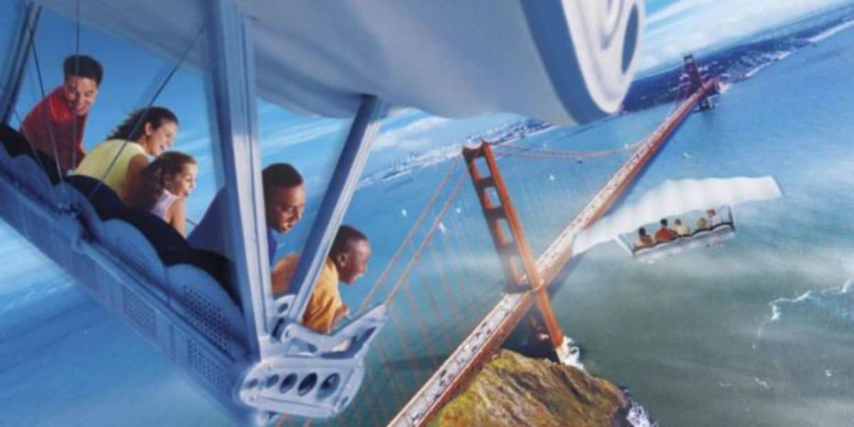 A group of people on the Soarin' ride at EPCOT simulates flying over the Golden Gate Bridge, with passengers smiling and leaning forward. The scene includes a blue sky and ocean below, capturing all the exhilaration of this aerial adventure.