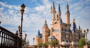 A fairytale-like castle with blue-topped towers and ornate spires stands against a partly cloudy sky, reminiscent of the whimsical charm of Zootopia. Decorative street lamps and a fence are visible in the foreground, nestled amid lush greenery.