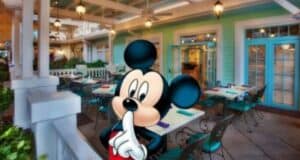 A cartoon character stands in front of Disney's Olivia's Cafe at Old Key West, a quaint outdoor eatery with teal chairs and tables adorned with colorful napkins. The building boasts a light green facade with blue trim, while hanging lanterns cast a warm glow.