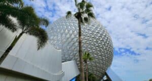 A large, geodesic sphere with a silver, triangular panel exterior stands timelessly against a partly cloudy sky. Palm trees line the foreground while a white, modern building curves gracefully on the left—a serene scene undisturbed by any tragedy at Disney World.