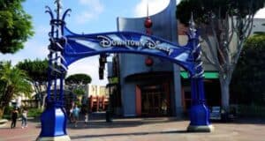 The entrance archway to Downtown Disney at Disneyland Resort showcases a blue and silver geometric design, surrounded by lush trees and bustling visitors in the background, all under a clear sky.