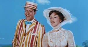 A smiling man and woman stand together against a blue sky with white clouds. The man wears a colorful striped suit, a bow tie, and a straw boater hat. The woman is dressed in a high-neck white dress with lace details and a wide-brimmed hat.