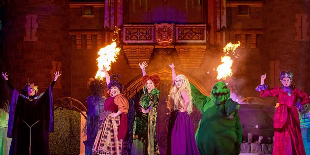 Disney villains perform on stage with dramatic gestures, flanked by flames, in front of a castle backdrop during a nighttime theatrical show.