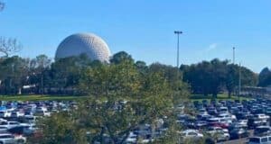 A bustling parking lot with numerous vehicles, with the iconic EPCOT geodesic sphere visible in the background against a clear blue sky.