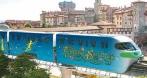A vibrant, decorated train featuring "fantasy springs" theme, with silhouettes of fairytale characters, travels on an elevated track through a scenic area with european-style buildings in the background.