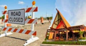 A split image showing road construction on the left with an orange and white "Road Closed" sign, and a building with Polynesian architectural design reminiscent of Disney's resort style on the right, featuring an A-frame structure and vibrant decorations.