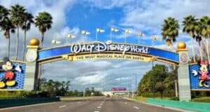 Entrance sign of Walt Disney World, featuring large bold letters and images of Mickey and Minnie Mouse, under a sunny sky with fluffy clouds, viewed from a road lined with palm trees near Typhoon