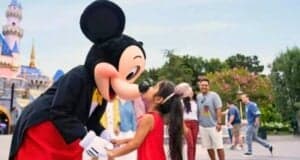 A girl in a red dress happily kisses Mickey Mouse on the nose at Disney, with other visitors and the castle blurred in the background.