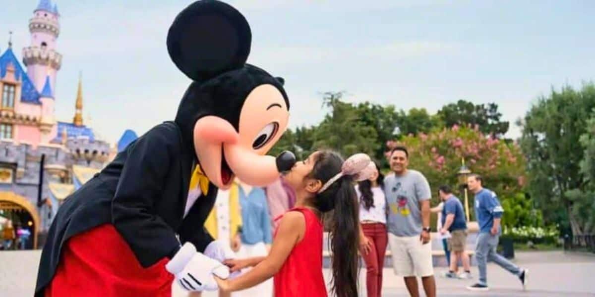 A girl in a red dress happily kisses Mickey Mouse on the nose at Disney, with other visitors and the castle blurred in the background.