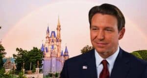 A man in a blue suit with a red tie stands superimposed on an image of disneyland featuring sleeping beauty's castle under a bright rainbow.