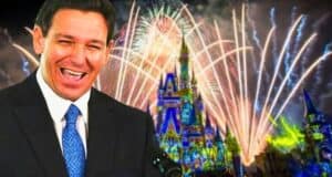 A man in a suit and tie is smiling broadly in front of a colorful night scene featuring a lit-up castle and fireworks in the sky. The castle has intricate lighting and projections, creating a celebratory atmosphere.