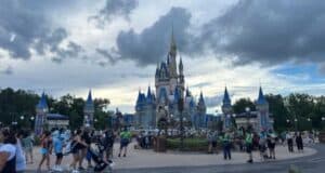 A bustling crowd gathers in front of a large, ornate castle with blue spires and golden accents under a partly cloudy sky at an amusement park. Paths are lined with lampposts and foliage, with visitors walking, pushing strollers, and taking photos.