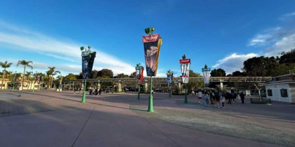 An entrance area to an amusement park with banners featuring various characters and themes hanging from green lampposts. Visitors gather near the entrance on a clear, sunny day with a few clouds in the sky. Trees and buildings are visible in the background.