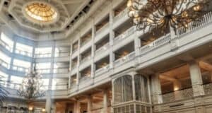 An opulent hotel atrium with a multi-story layout, featuring balconies lined with white railings. A grand chandelier hangs centrally, and sunlight streams from a large, ornate dome at the top, illuminating the space with a warm glow.