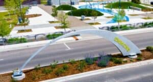 A modern public park featuring a large arched structure with the word "EMMONS" on a street median. The park has curved pathways, grassy mounds, young trees, and landscaped areas with purple flowers. The scene is vibrant and well-maintained.