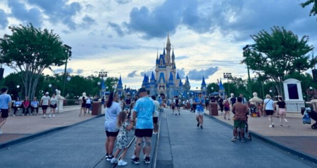 Visitors walk down a broad pathway towards a grand, iconic castle with blue and gold accents. The sky is partly cloudy, and trees line the pathway. People are taking photos, chatting, and enjoying the theme park atmosphere.