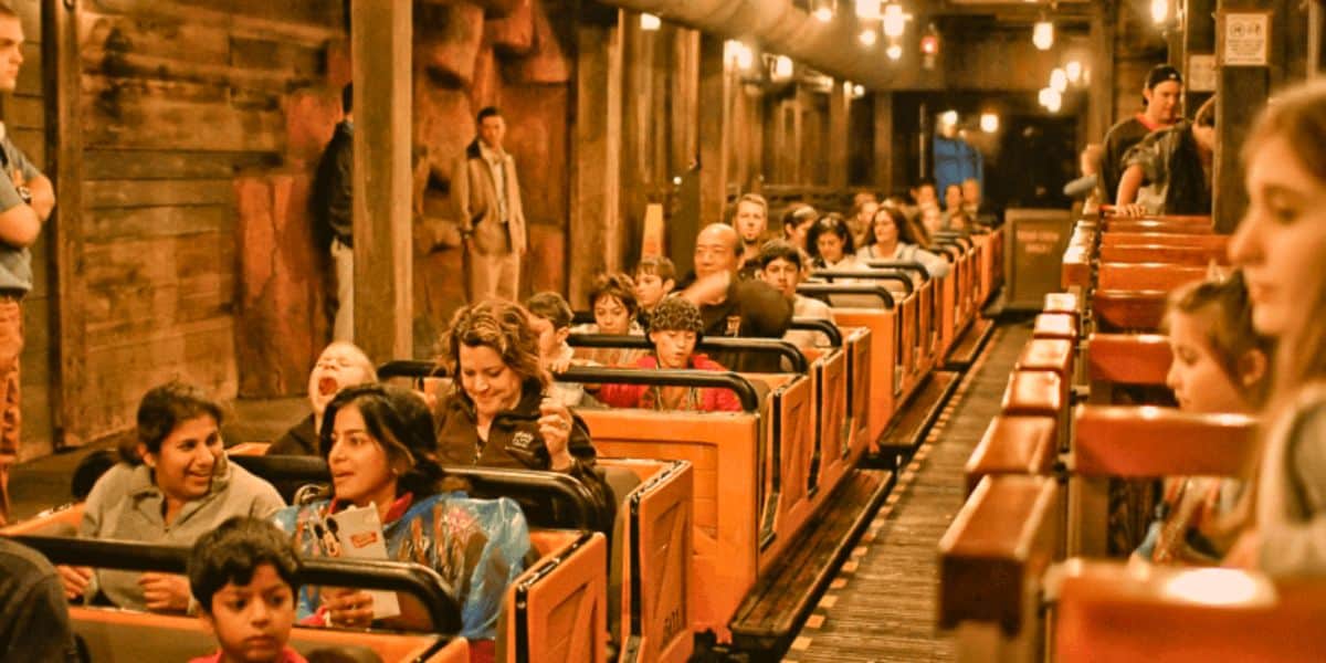 People sitting in a themed amusement park ride vehicle appear excited and engaged, with dim, warm lighting enhancing the rustic setting as they prepare for the ride to start.