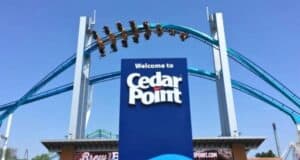 A large blue sign at the entrance of Cedar Point amusement park with the text "Welcome to Cedar Point" in white. Above the sign, people are experiencing an exhilarating roller coaster ride, soaring through the air with the blue track in clear view against a bright sky as PETA advocates nearby.