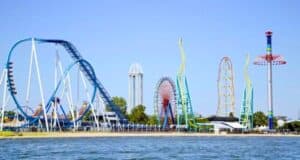Image of Cedar Point amusement park with large roller coasters, a Ferris wheel, and a tall drop tower. The park is situated beside a body of water, with clear blue skies overhead. The vibrant rides and structures stand out against the green trees and open space.